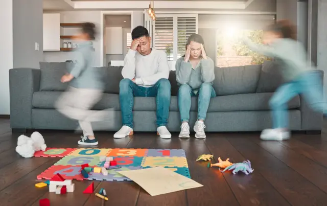 Dois pais jovens parecendo cansados sentados no sofá, enquanto seus filhos correm pela casa
